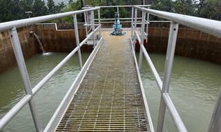 A walkway of grates leads over a greenish water surface, flanked by industrial structures and equipment in a natural setting.