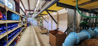 A spacious industrial corridor featuring storage shelves, blue bins, wooden crates, and green pipes alongside a large blue valve.