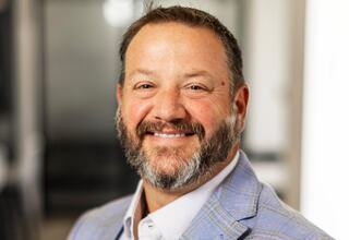 Shane Harris, Principal at FGM Architects; Smiling man in a light blue blazer with a beard, posed in a well-lit, modern setting. Professional and approachable demeanor.