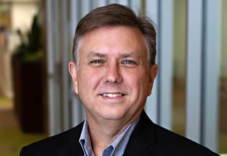 Steve Hafer, Principal and Vice President at FGM Architects; A man with short gray hair and a friendly smile wears a dark blazer over a checked shirt, standing against a blurred office background.