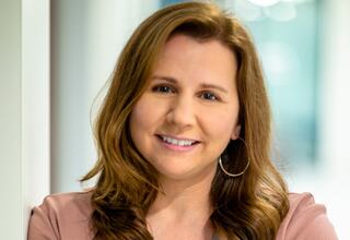 Portrait of Jen Carlson, Architect and Project Manager at FGM Architects; A woman with long, wavy brown hair and a warm smile wears a pink blazer. She stands in a bright, modern setting.