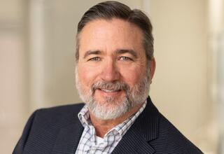 Principal Bryan Mason at FGM Architects; A smiling man with a well-groomed beard, wearing a navy blazer over a checked shirt, poses confidently against a blurred background.