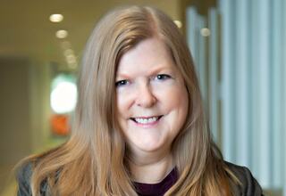 Rebecca Richter, Architect at FGM Architects; A smiling individual with long, straight hair poses in a softly lit, modern space, exuding warmth and approachability.