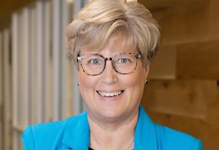 Kris Waugh, PhD, Educational Researcher and Planner at FGM Architects; A smiling professional with short, blonde hair, wearing glasses and a bright blue blazer, stands in a well-lit, modern setting.
