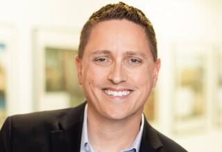 Troy Kerr, Vice President and Principal at FGM Architects; A man with a friendly smile, wearing a blazer over a collared shirt, stands in a well-lit space with framed pictures in the background.