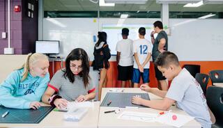 Students collaborate at desks while others observe a presentation on a screen, fostering engagement and teamwork in an educational setting.