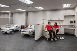 A modern healthcare simulation room features two individuals seated together, with hospital beds and medical equipment in the background.