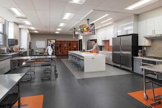 A modern kitchen classroom features sleek appliances, a large central island, and organized storage, with two individuals preparing food.