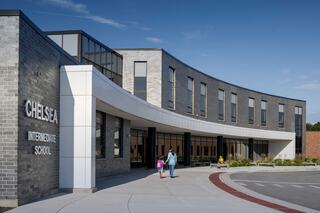 A modern school building features a curved façade, large windows, and students walking towards the entrance on a sunny day.