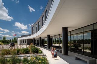 Two children walk along a curved entrance of a modern building, surrounded by greenery and bright blue sky, with a flag visible nearby.