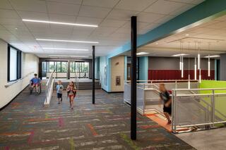 Bright, modern school interior featuring colorful flooring, groups of children running, and open spaces with stairways and lockers visible.