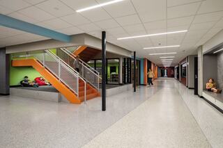 A modern hallway features colorful walls, students lounging, and an orange staircase leading to upper levels, creating a vibrant atmosphere.
