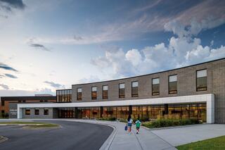 A modern brick building with large windows and a curved entrance, set against a dramatic sky, features two children walking towards it.
