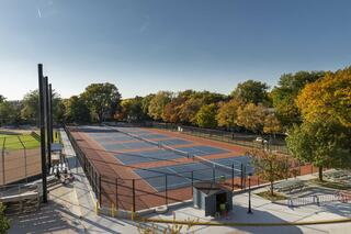 A spacious outdoor tennis complex features several courts amidst vibrant autumn foliage under a clear blue sky. Activity fills the area.
