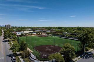 A lush sports complex features a soccer field and baseball diamond, surrounded by trees and urban views in the background.