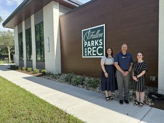 Three individuals pose outside a modern building with a sign reading "O'Fallon Parks and Rec," surrounded by landscaping and a walkway.