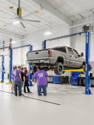 A group of people watches as a silver truck is lifted by a hydraulic lift in a bright, spacious automotive workshop.