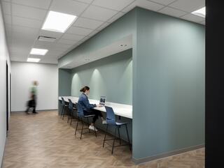 A modern workspace features a woman at a desk on the right, while a person walks by in a hallway with a light color scheme and wood flooring.