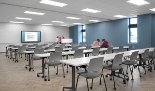 A modern classroom features rows of tables and chairs, a display screen, and students engaged in collaborative learning.