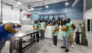 A group of trainees observes a welder working in a bright, modern workshop, all wearing protective gear for safety.