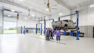 A group of people in purple shirts gathers around a lifted truck in a spacious automotive workshop with bright lighting and clean floors.