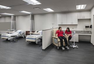A modern healthcare setting features two individuals in red shirts, seated together, with hospital beds and medical equipment in the background.