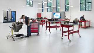 A modern workshop features multiple workstations, mechanics assembling engines, and an instructional screen. Bright and spacious environment.