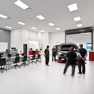 A modern automotive workshop features technicians gathered around a vehicle on a lift, with workstations and equipment in the background.