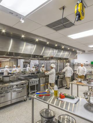 A modern culinary lab filled with student chefs in white uniforms, actively cooking and preparing various dishes amid stainless steel appliances.