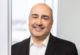 A smiling man in a business suit poses confidently near a window, offering a welcoming and professional demeanor.