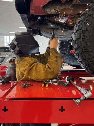 A welder, wearing protective gear, works on a vehicle's undercarriage, sparks flying in a well-lit garage filled with tools and cars.