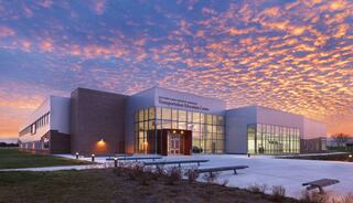A modern educational building with large glass windows stands against a vibrant sunset, highlighting its contemporary architecture and surrounding greenery.