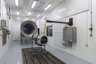 A clean room features a jet engine on a stand, a large exhaust outlet, control panel, and bright overhead lighting, emphasizing industrial precision.
