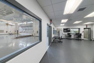 A modern, clean control room with large interior windows, computers, and a viewing area leading to a spacious industrial section.