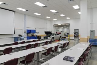 A spacious automotive classroom features workstations, chairs, and vehicles, with natural light and a projector screen for demonstrations.