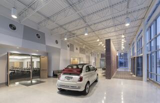 A modern, spacious lobby features a white car parked beside a brick wall, with high ceilings and large windows allowing natural light.