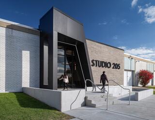 A modern building features a striking black entrance, with two individuals approaching via a concrete path and lush greenery surrounding it.