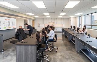 A modern hairdressing classroom with students observing techniques on a model, featuring sleek workstations and instructional displays.