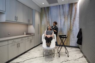A woman receives a treatment in a modern room featuring sleek cabinetry, marble flooring, and a serene water-themed backdrop.