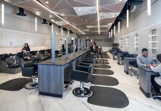 A modern salon features sleek styling stations, black chairs, and bright lighting, with clients receiving services in a spacious layout.