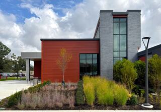 A modern building features a mix of gray brick and orange siding, surrounded by vibrant landscaped plants and a clear blue sky.