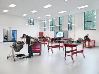 A bright workshop filled with red workbenches and automotive engines, where individuals are engaged in hands-on tasks and learning.