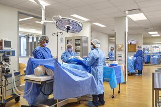 Medical trainees in blue scrubs engage in a mock surgical procedure in a well-equipped operating room. Hospital environment is visible.