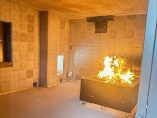 A stark room with concrete walls features a prominent fire burning in a black grill, creating an intense, dramatic atmosphere.