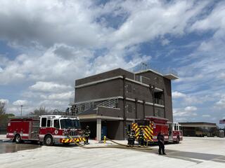 Firefighters train on a modern facility with two fire trucks, hoses laid out, and dramatic clouds overhead. Safety and skill in action.