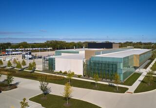 A modern commercial building features large glass walls, surrounded by lush landscaping and ample parking under a clear blue sky.