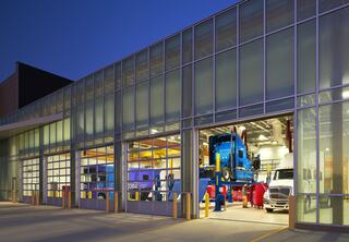 A modern repair shop at dusk, featuring large glass doors, a blue truck on a lift, and various vehicles inside waiting for service.