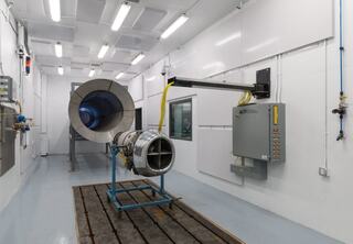 A clean, industrial testing facility featuring a jet engine mounted on a stand, with a large test chamber in the background.