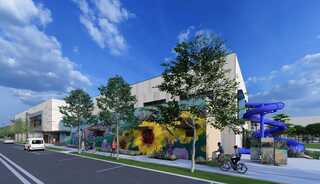 A modern building features colorful murals, green landscaping, and a vibrant blue water slide, set against a bright, cloudy sky.