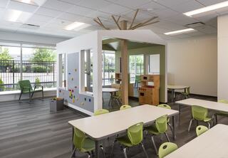 A bright, modern classroom featuring a playhouse with a tree design, small tables, and green chairs.